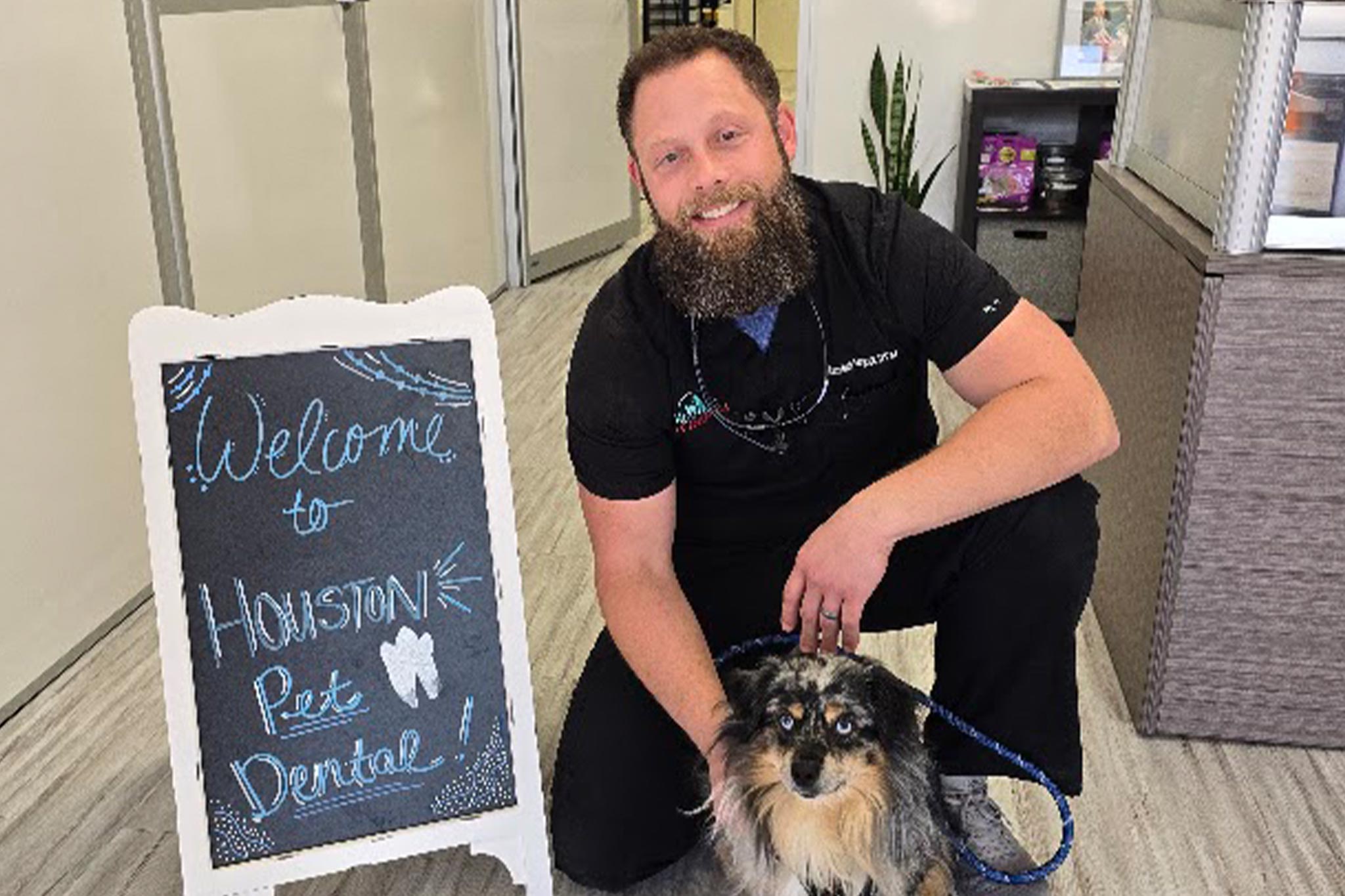 male veterinarian smiling while petting small fluffy dog in front of chalkboard sign that says "Welcome to Houston Pet Dental!"